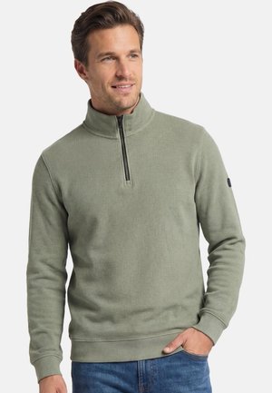 Man draagt een lichtgroene pullover met kwartrits en opstaande kraag en een blauwe spijkerbroek, glimlacht en kijkt iets naar rechts, hand in de zak.