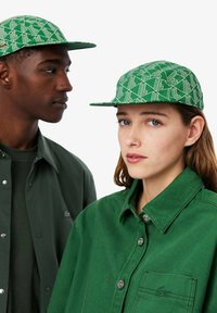 Il cappello verde a fantasia presenta una visiera piatta e un sottile design geometrico. I modelli indossano camicie verdi abbinate, che mettono in mostra colori solidi e texture.