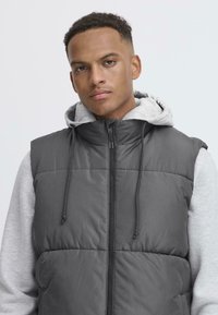 Grå puffy vest med glidelås foran, med hette i lys grå. Vestgen har en quiltet tekstur og justerbare snorer rundt halsen.