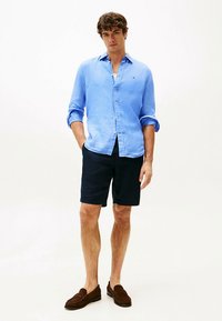 Camisa de linho azul-claro com mangas arregaçadas, calções azul-marinho escuro e mocassins castanhos, em pose de pé contra um fundo neutro.