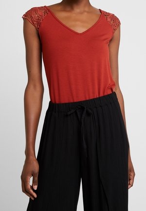 Kobieta w rdzawo-czerwonym topie z dekoltem w serek, z koronkowymi krótkimi rękawkami typu cap oraz luźnych czarnych spodniach z sznurkiem, stojąca na jednolitym tle.