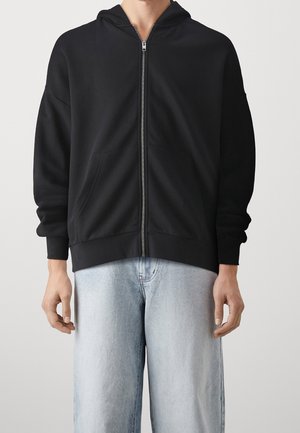 Black zip-up hoodie, soft fabric, oversized fit, kangaroo pockets, ribbed cuffs and hem, paired with light blue jeans.