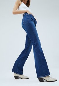Blaue, auslaufende Jeans aus Denim mit hoher Taille, ausgestattet mit einem klassischen Fünf-Taschen-Design und sichtbaren Nähten. Getragen mit beigefarbenen, hohen Ankle Boots.