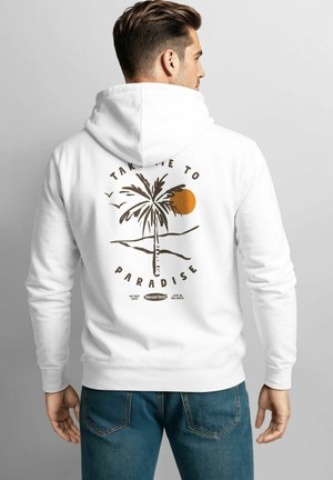 Mann mit weißem Kapuzenpullover, auf dem eine Palme, eine orangefarbene Sonne, Vögel und der Text "Bring mich ins Paradies" auf dem Rücken gedruckt sind.