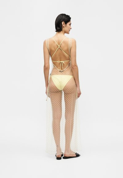 Γυναίκα με κοντά σκούρα μαλλιά που φοράει διαφανές πουά φόρεμα πάνω από κίτρινο κάτω μέρος μπικίνι και μαύρα σανδάλια τύπου slide, με το πρόσωπο στραμμένο προς τα πίσω σε λευκό φόντο.