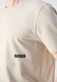 Beige bomullströja med rund hals och korta ärmar, med en svart "BOSS"-logo broderad på vänster bröst.