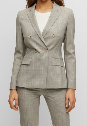 Kvinde iført beige ternet dobbeltradet blazer med matchende bukser og en hvid skjorte under, stående med afslappede arme.