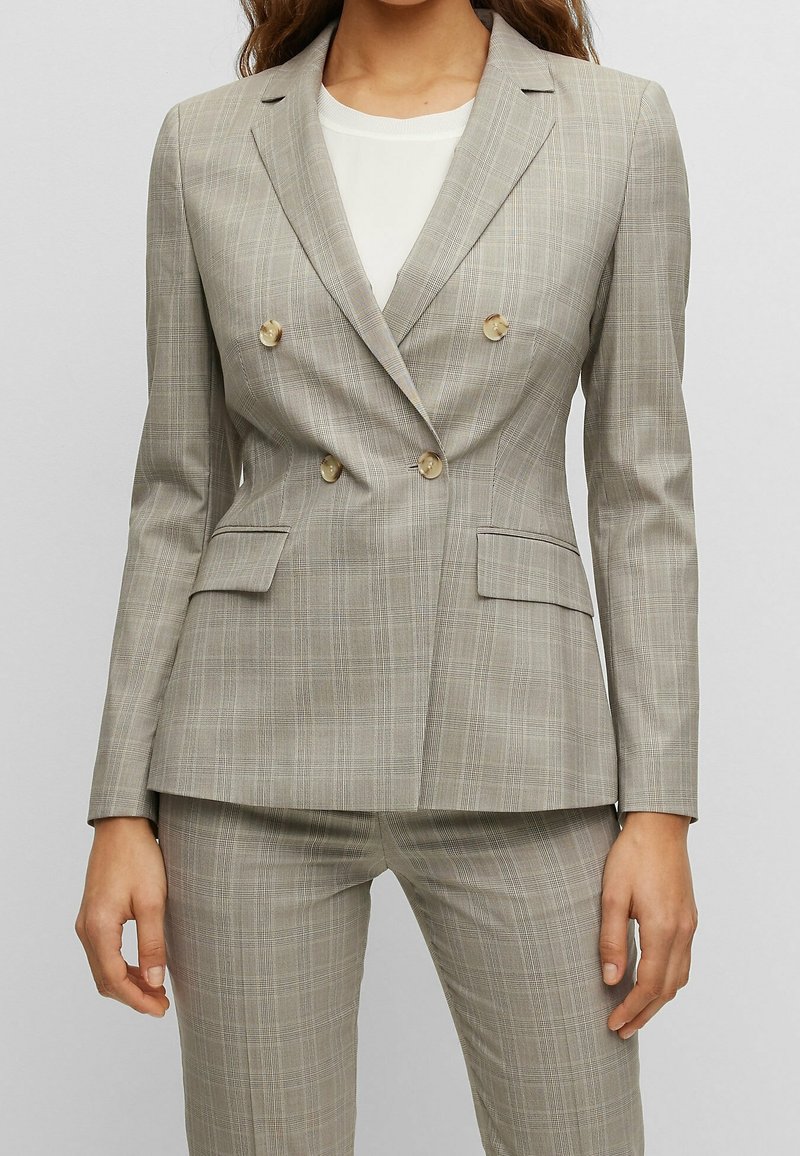 Femme portant un blazer croisé à carreaux beige avec un pantalon assorti et une chemise blanche en dessous, debout avec les bras détendus.