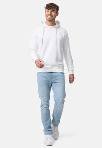Hvid hættejakke med snore og frontkangaroo-lomme, parret med lyseblå jeans og hvide sneakers, båret af en mand der går.