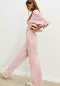 Žena v bledě růžových širokých kalhotách a odpovídajícím oversized topu, která napíná jednu nohu vpřed proti bílé zdi a béžové podlaze.