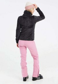Zwarte fleece jack met hoge kraag, gecombineerd met lichtroze broek met zijritsen. Het model draagt een witte en roze beanie.