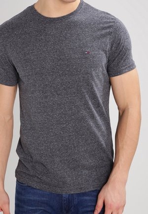 Man die een effen donkergrijs, korte mouw, T-shirt met ronde hals en een blauwe spijkerbroek draagt, staand met één hand ontspannen naast zich.