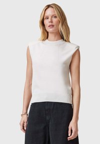 Mouwloze, crèmekleurige gebreide top met ribbelstructuur bij de hals en zoom. Heeft subtiele verticale stiksels en wordt gecombineerd met donkere stoffen broek.