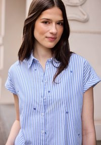 Jeune femme aux longs cheveux foncés portant une chemise boutonnée à rayures verticales bleues et blanches avec de petits cœurs brodés.