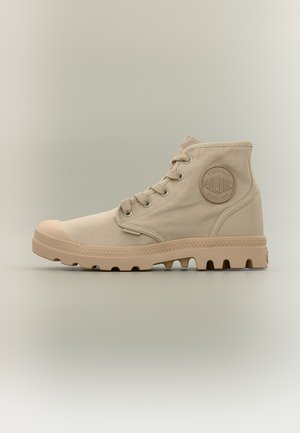 Bota alta de lona beige con suela de goma, puntera redonda, ojales metálicos, cordones y parche circular de la marca en el tobillo.