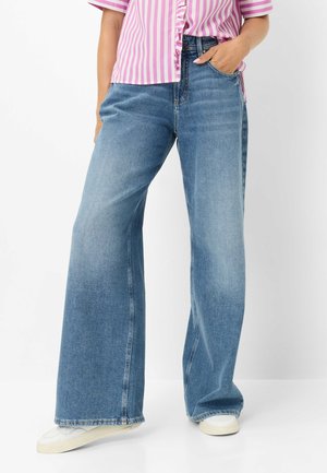Person iført blå jeans med vide ben, hvide sneakers og en lyserød og hvid stribet kortærmet skjorte med den ene hånd i lommen.