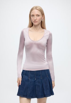 Donna bionda che indossa un top aderente a maniche lunghe rosa pallido e una gonna plissettata di jeans blu scuro, in piedi su sfondo bianco.