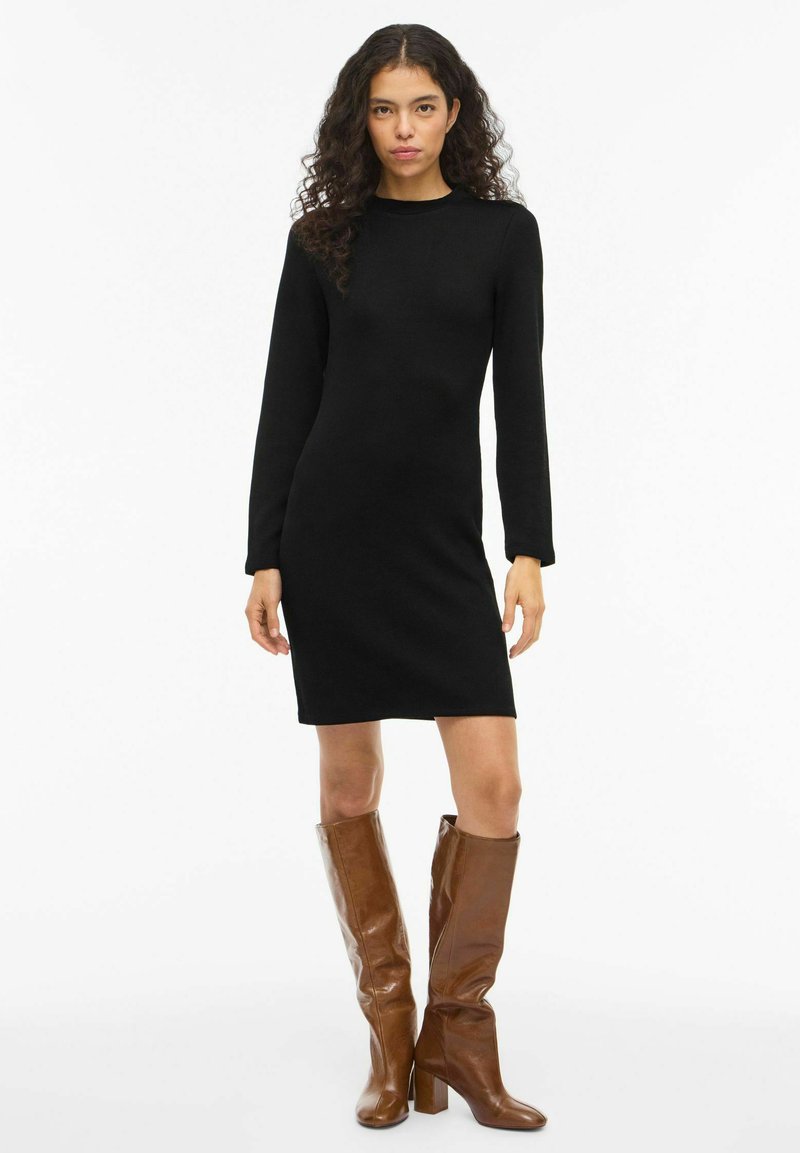 Czarna sukienka dzianinowa z długim rękawem o prostej sylwetce. Połączona z brązowymi skórzanymi kozakami na obcasie sięgającymi do kolan. Prosty design, bez wzorów.