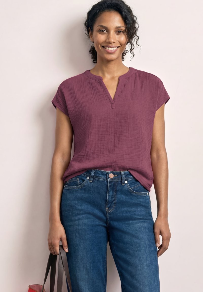 Femme souriante aux cheveux bouclés portant un chemisier violet texturé à manches courtes et un jean bleu, tenant un sac à main marron foncé à ses côtés.