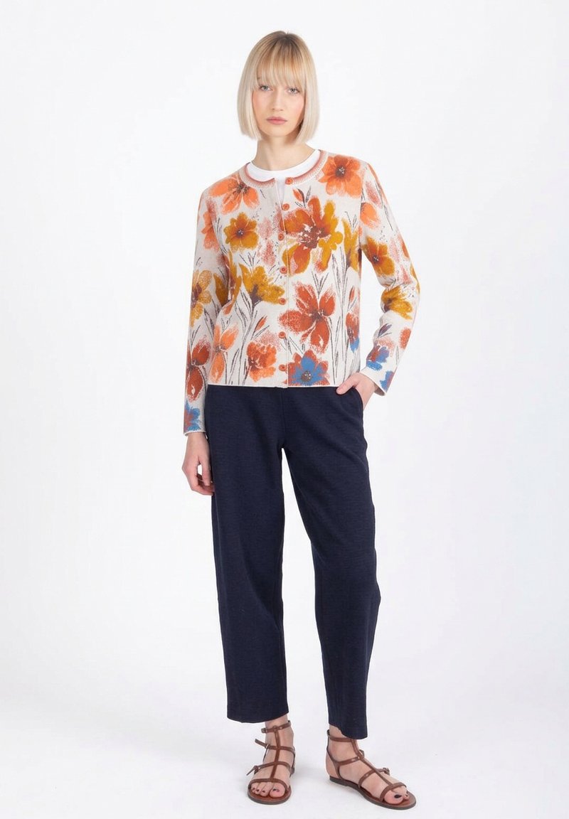 Ženska, oblečena v večbarvni cvetlični kardigan, mornarsko modre hlače in rjave sandale s trakovi, stoji z eno roko v žepu.