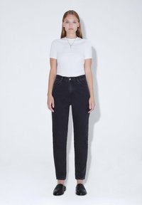 Schwarze hochgeschnittene Jeans mit schmal zulaufendem Bein, kombiniert mit einem taillierten weißen Kurzarmshirt. Glatter Baumwollstoff; minimalistische Details.