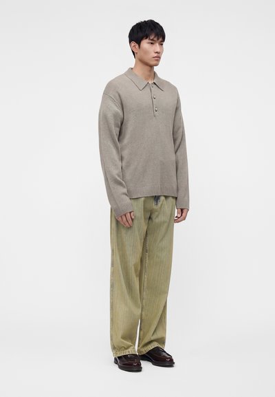 Homme debout portant un pull en maille beige boutonné, un pantalon large en velours côtelé vert olive et des chaussures en cuir marron foncé sur un fond uni.