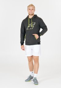 Sudadera gris con detalles en amarillo neón y diseño gráfico, combinada con pantalones cortos blancos y zapatillas deportivas grises, de pie sobre un fondo sencillo.
