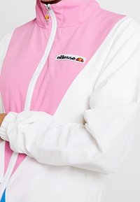 Biała i różowa kurtka przeciwwiatrowa z wysokim kołnierzem, pełnym zamkiem z przodu i elastycznymi mankietami. Posiada logo na lewej piersi oraz wzór w kolorowych blokach.
