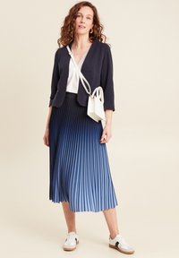 Blazer blu navy sopra una maglietta bianca, abbinato a una gonna plissè con gradiente che va dal blu scuro al blu chiaro. Una borsa bianca e sneakers casual completano l'outfit.