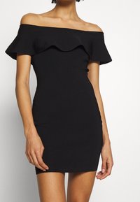 Abito nero scollato sulle spalle con scollo a balze. Realizzato in tessuto a coste, presenta una silhouette aderente e una lunghezza mini.