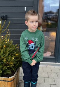 Grön stickad tröja med en björn som snowboardar med en röd bräda, snöflinge-mönster, bärs över mörka byxor och blå gummistövlar.