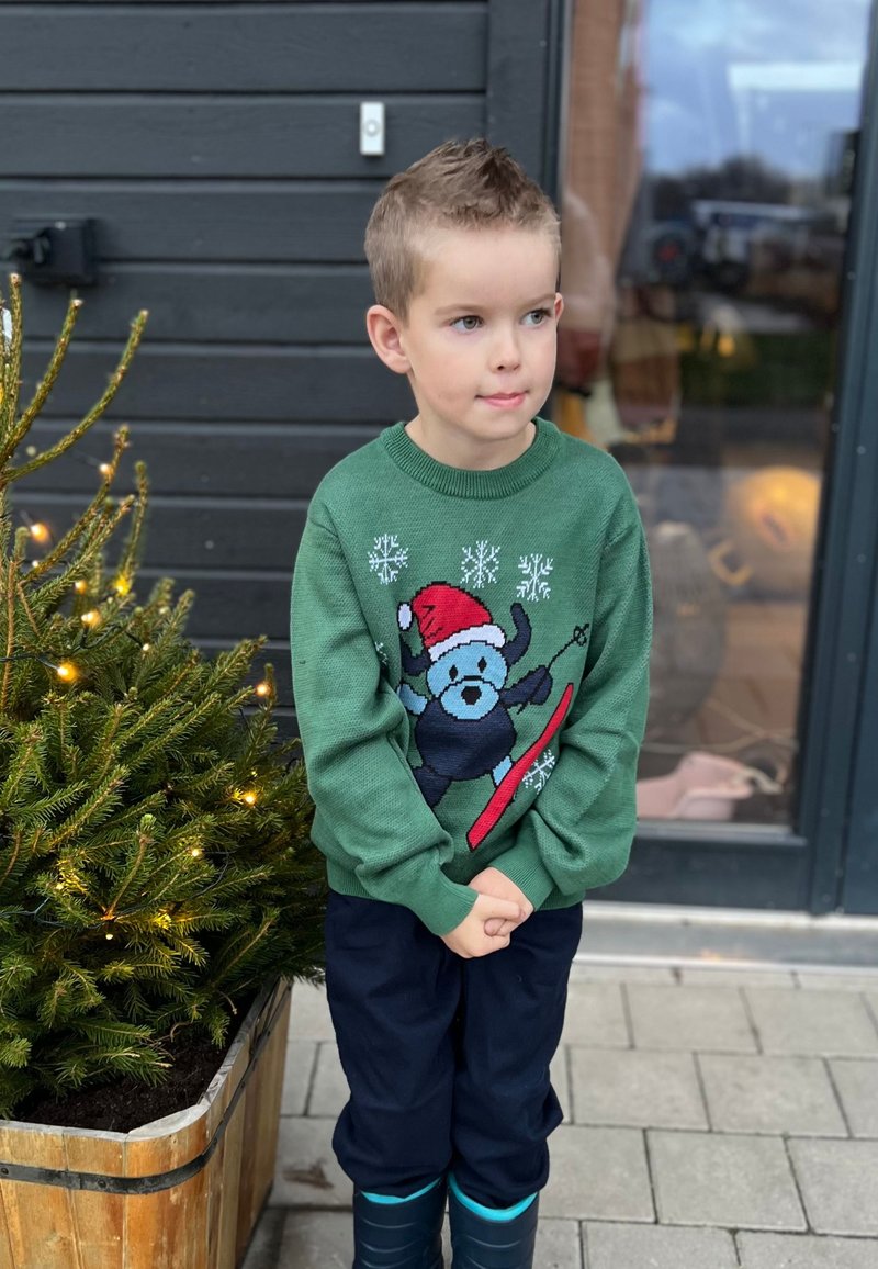 Grön stickad tröja med en björn som snowboardar med en röd bräda, snöflinge-mönster, bärs över mörka byxor och blå gummistövlar.