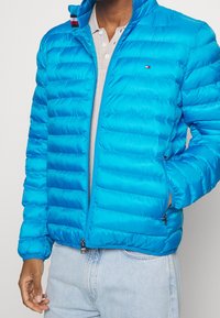 Giubbotto puffer blu con design a caramella orizzontale, chiusura con zip e colletto alto. Presenta un dettaglio del logo e due tasche laterali.