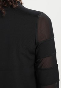 Mustapaita, jossa on läpikuultavat hihnat ja vaakasuorat kangaspaneelit. Sujuva kangas, mukautuva malli ja pyöreä kaula-aukko.