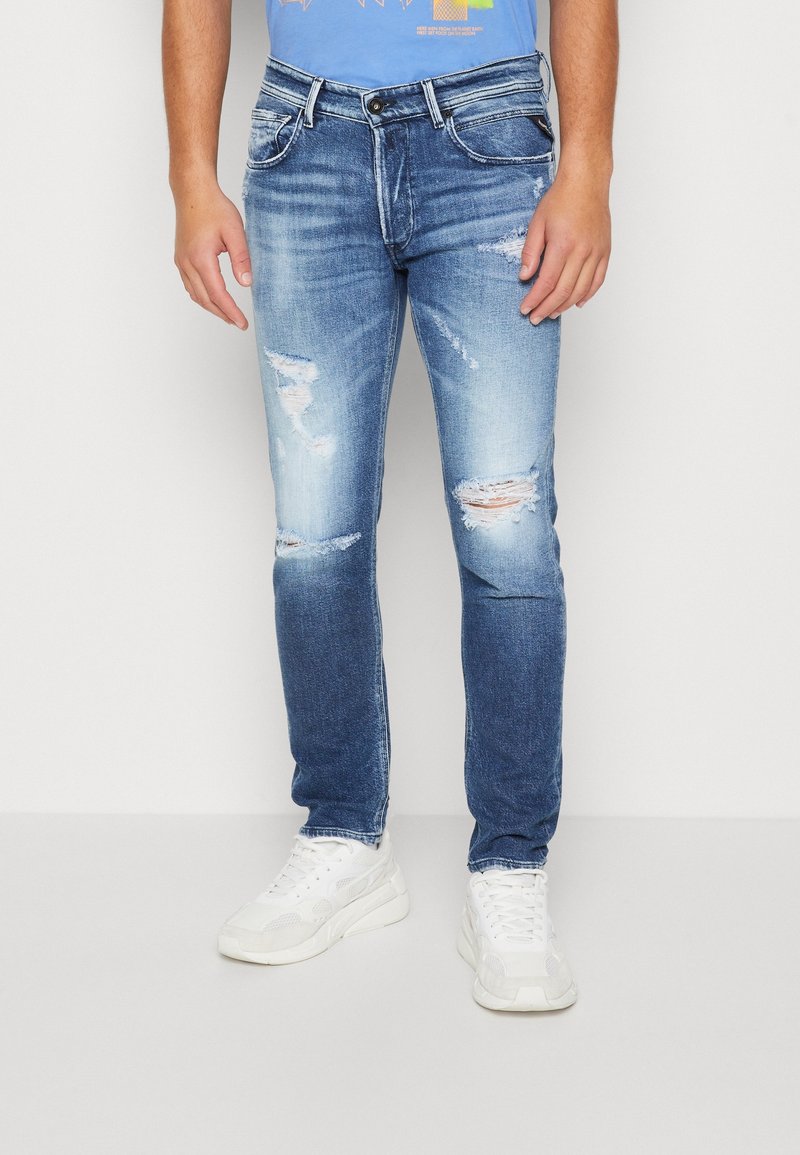 Hombre vestido con jeans ajustados de mezclilla azul claro desgastados y zapatillas blancas, de pie contra un fondo blanco liso.