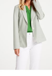 Blazer rayé vert clair avec un col à revers, deux boutons et des poches frontales, associé à un haut vert et un pantalon blanc.