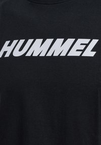 Camiseta negra con texto blanco en mayúsculas y negrita "HUMMEL" centrado en el pecho, mostrando parte del escote y la textura de la tela.