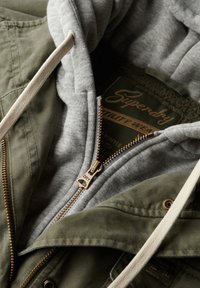 Chaqueta verde oliva con capucha y forro gris, que presenta un cierre dorado, cordones y una etiqueta en el cuello interno. Tejido texturizado.