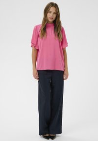 Blusa rosa con mangas abullonadas, cuello alto y textura suave. Combinada con pantalones de pierna ancha en navy y bailarinas negras de punta.