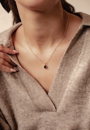 Goldene Halskette mit einem tropfenförmigen grünen Edelsteinhänger. Über einem beigen, strukturierten Pullover mit tiefem V-Ausschnitt getragen. Glatte, polierte Details.