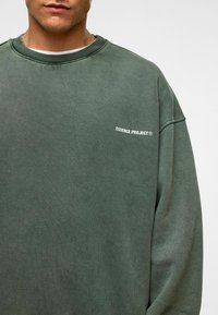Homme portant un sweatshirt à col rond vert délavé avec le texte "ESSENCE PROJECT®" sur la poitrine gauche, devant un fond uni.