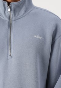 Light blue fleece pullover with a high neckline, half zip, and embroidered "Hollister" logo on the left chest. Soft texture and casual fit.