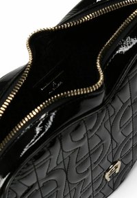 Bolso de cuero negro brillante con diseño acolchado, cremallera dorada y una pequeña etiqueta con el logo en el interior. El interior está forrado con una tela negra suave.