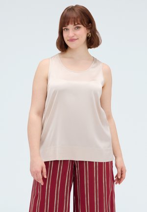 Junge Frau mit kurzen braunen Haaren, die ein ärmelloses beiges Satintop und rot gestreifte weite Hosen trägt, steht vor einem schlichten Hintergrund.