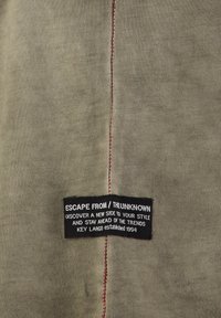 Tejido caqui con una textura suave, que presenta una costura roja prominente a lo largo de la costura y una etiqueta negra con texto blanco en la esquina inferior izquierda.