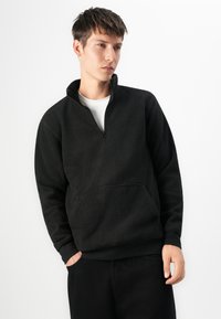 Joven vestido con un pulóver negro de media cremallera de forro polar y pantalones negros, de pie con una mano en el bolsillo frente a un fondo blanco.