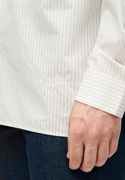 Κοντινό πλάνο ενός χεριού δίπλα σε ένα λευκό πουκάμισο μακρυμάνικο με λεπτές κάθετες ρίγες και κεντημένο κείμενο "maison kitsuné."