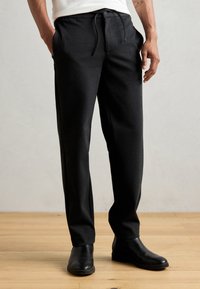 Pantalon noir fuselé en tissu doux avec taille élastique et poches latérales. Porté avec des bottines noires sur un sol en bois.