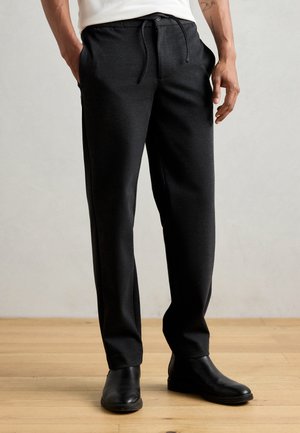 Pantalon noir fuselé en tissu doux avec taille élastique et poches latérales. Porté avec des bottines noires sur un sol en bois.
