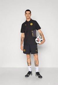 Čierne polo tričko s žltými akcentmi, s vreckom na zips a logom. Kombinované s čiernymi šortkami. Drží futbalovú loptu.
